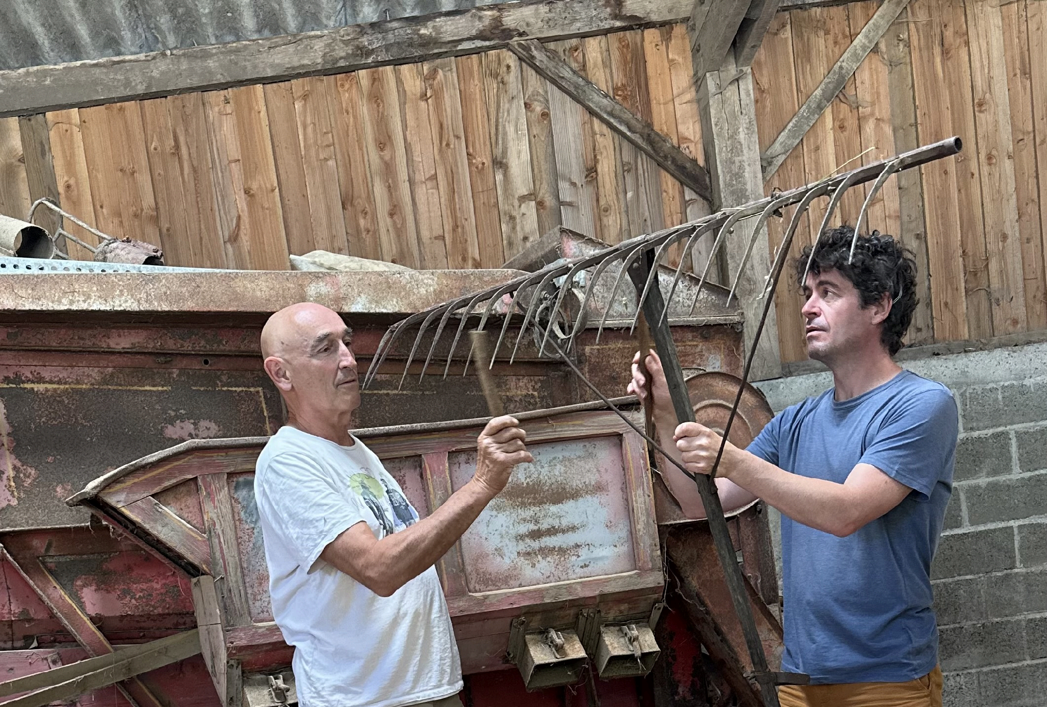 Benoît Sicat et Emmanuel Cury manipulant un râteau agricole ancien devant une machine agricole rouillée dans une grange